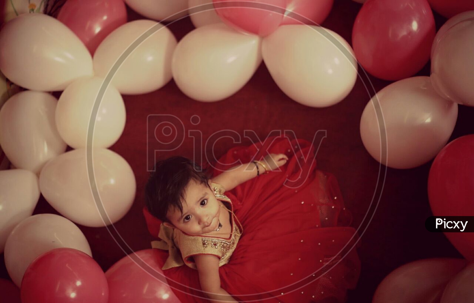 Indian baby Girl in red Frock Looking into Camera with Baloons All Around Her