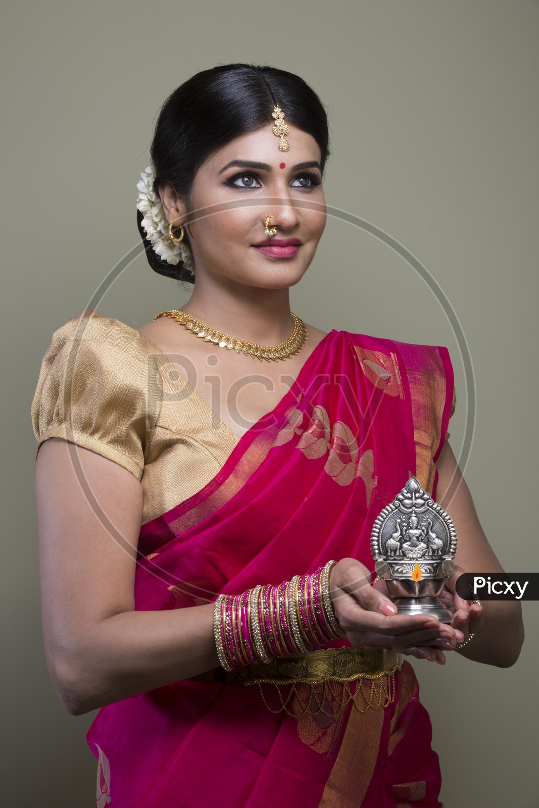 A Beautiful Indian Female Model  in Traditional Attire Wearing a Saree and Jewelry with an Expression and With a Traditional Silver Oil Lamp In Hands  on an Isolated Gery Background