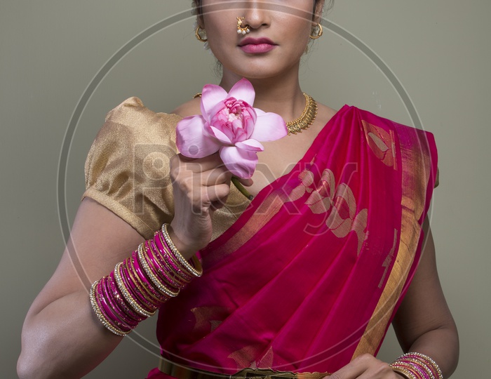 A Beautiful Indian Female Model  in Traditional Attire Wearing a Saree and Jewelry with an Expression and With a Lotus Flower in Hand  on an Studio Closeup shot