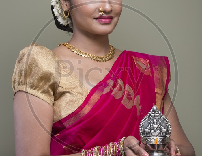 A Beautiful Indian Female Model  in Traditional Attire Wearing a Saree and Jewelry with an Expression and With a Traditional Silver Oil Lamp In Hands  on an Isolated Gery Background
