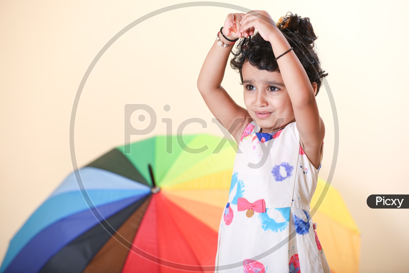 Indian girl dressed in colorful frock playing with her hair with rainbow colored umbrella in the background