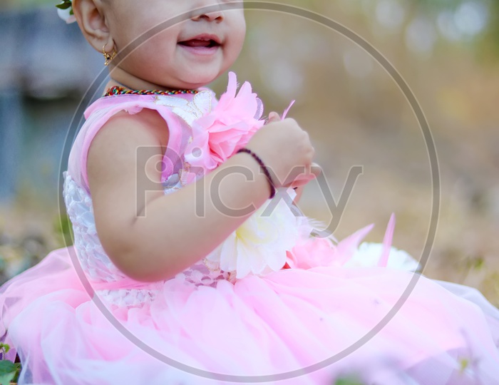 Baby girl in Pink frock with a flower hair band sitting in the middle of the fields and smiling