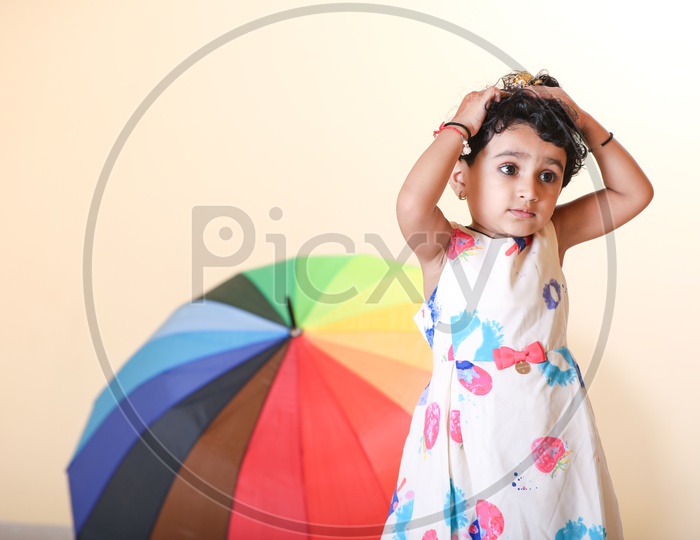 Indian girl dressed in colorful frock playing with her hair with rainbow colored umbrella in the background