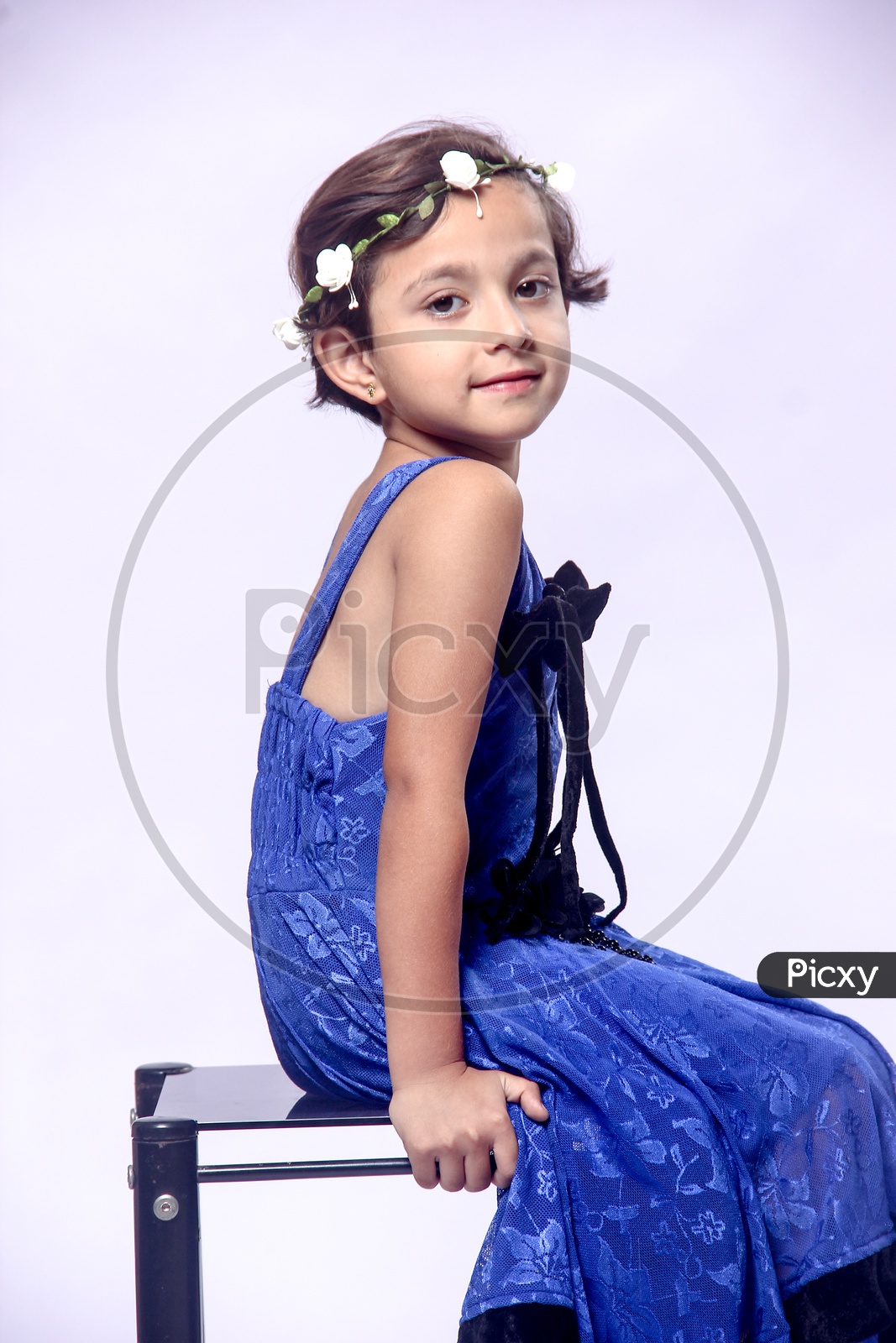 Girl dressed up in blue frock with a flower hair band, sitting on a teapoy and posing