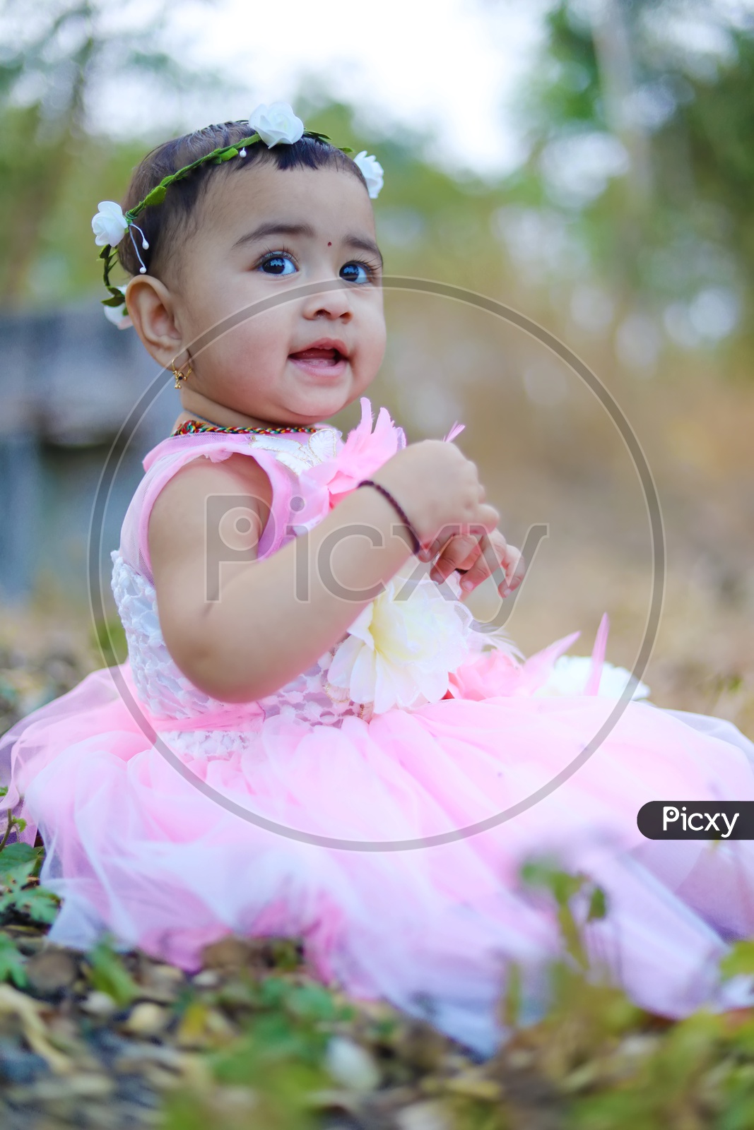Baby girl in Pink frock with a flower hair band sitting in the middle of the fields and smiling