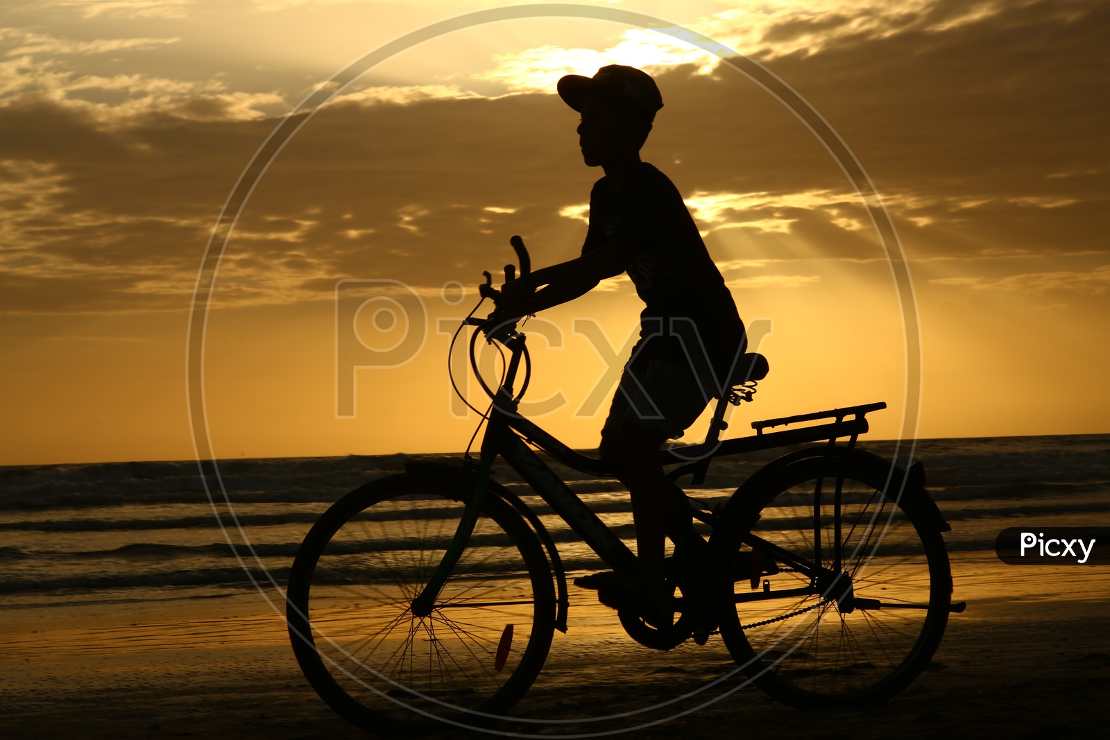 Young Boy Riding Bicycle Over a Golden Sunset Sky In Background At a Beach in Goa