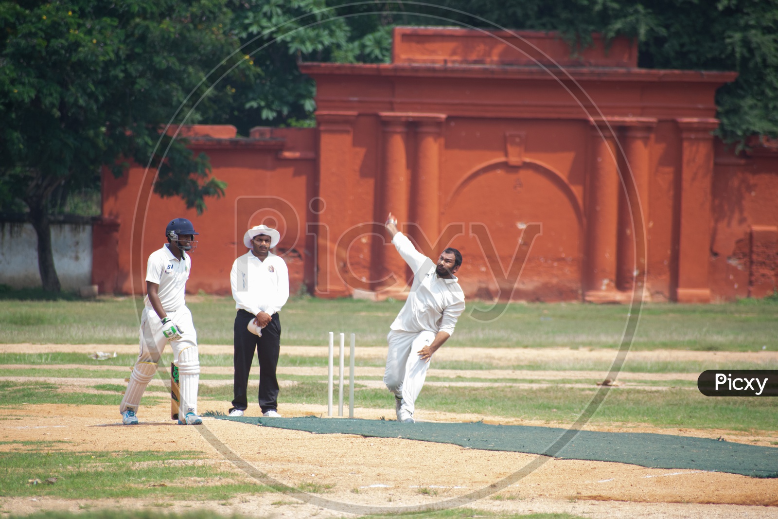 A bowler is bowling on the Cricket field while the empire watch and the batsman ready to take a run