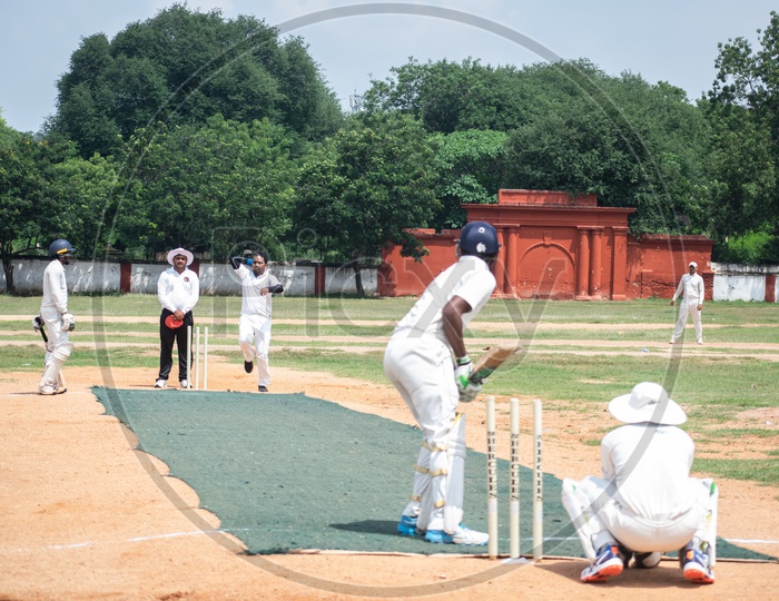 A bowler is bowling on the Cricket field while the empire watch and the batsman on the other end