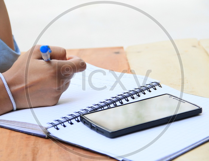 Image of Student Taking Notes In a Notebook And Smartphone on a Wooden ...