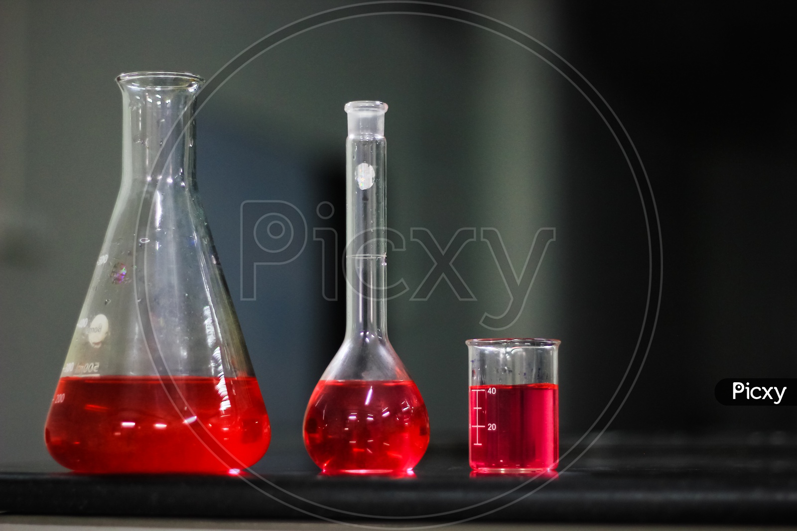 Red Liquid In A Round Bottomed Flask And Glass Beaker And Conical Flask On A Black Granite Table In Dark Background