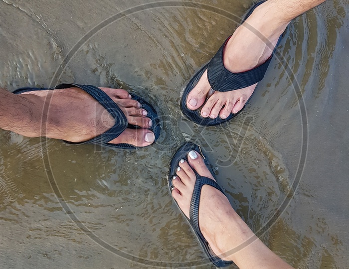 Three Tourists Getting Their Feet Wet With Slippers Together In Sea Water On Sandy Beach