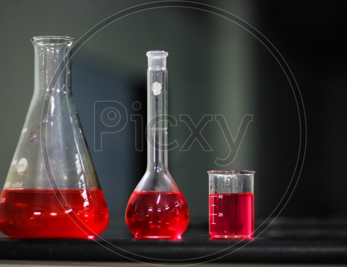 Red Liquid In A Round Bottomed Flask And Glass Beaker And Conical Flask On A Black Granite Table In Dark Background