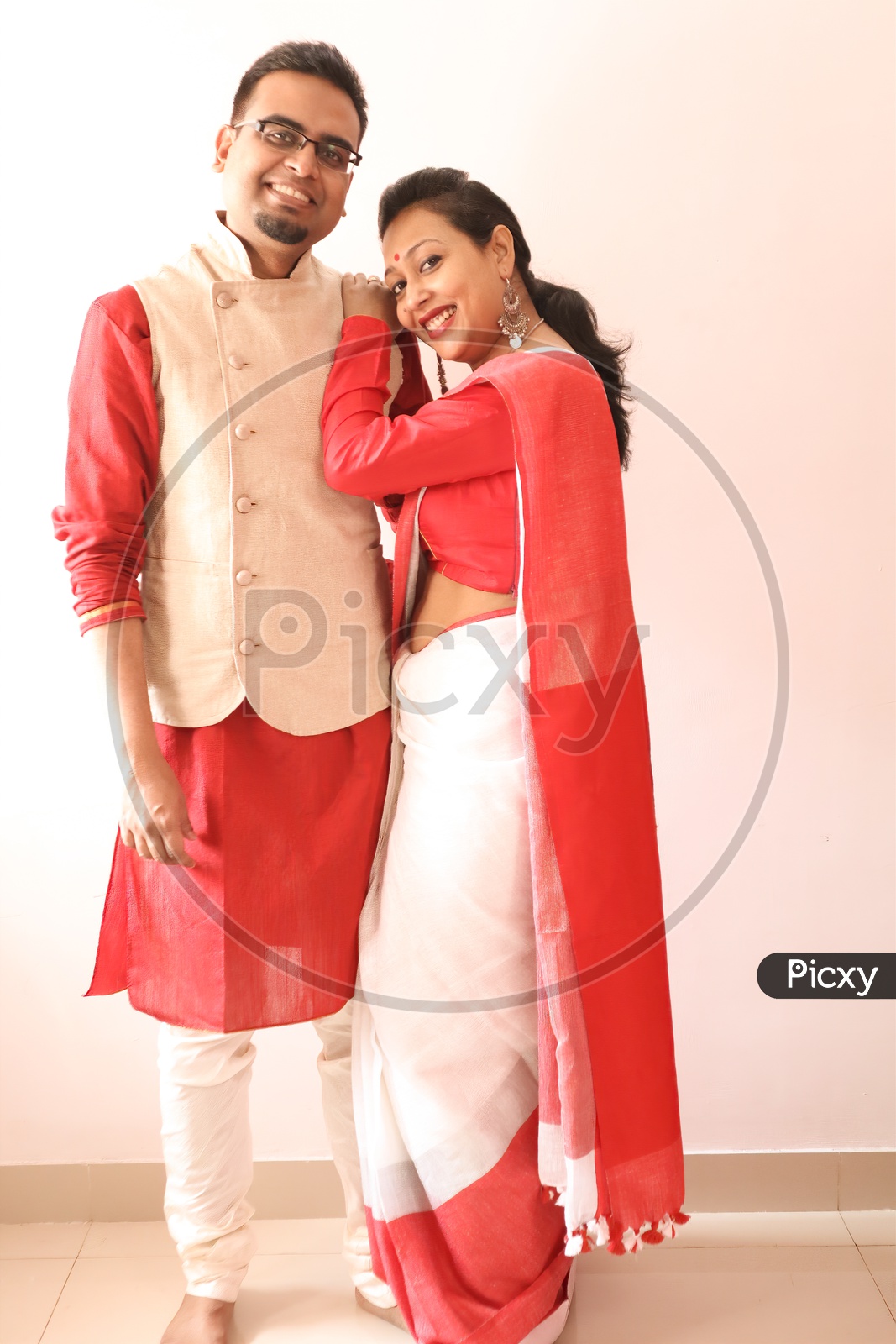 A Young Indian Bengali Assamese Married Romantic Couple Dressed In Red And White Ethnic Indian Dress, Looking At The Camera And Smiling