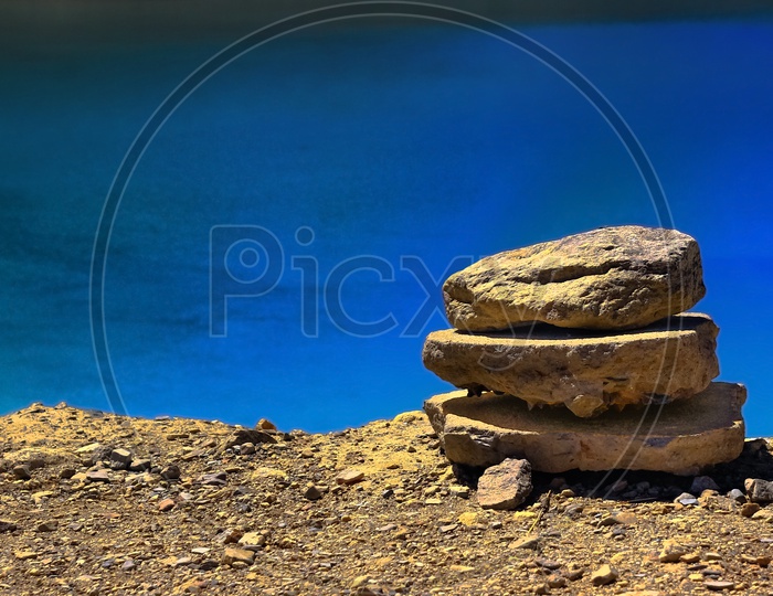 Heap Of Pebble Stacked On Lake Shore With Still Blue Waters In The Background