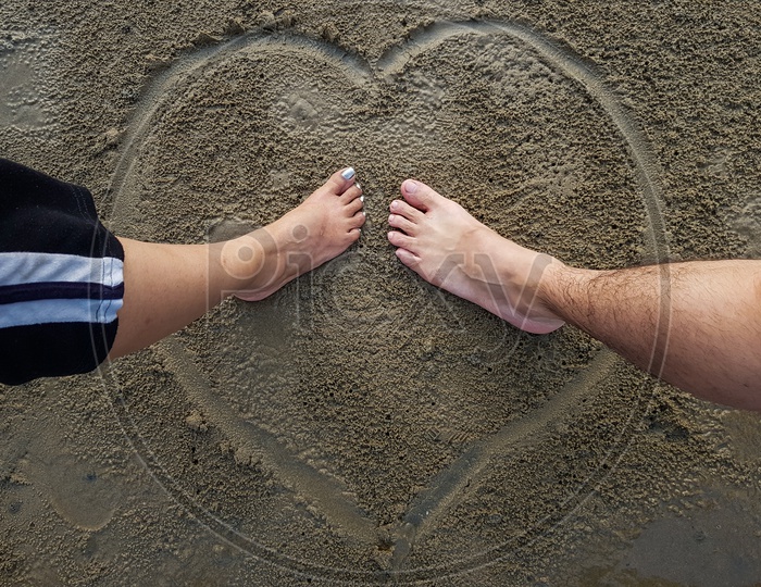 Romantic Couple Putting Feet Together On Heart Sign Background Drawn On Wet Beach Sand