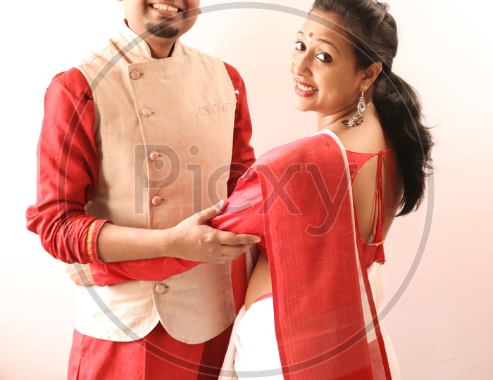 A Young Indian Bengali Assamese Married Romantic Couple Dressed In Red And White Ethnic Indian Dress, Looking At The Camera And Smiling