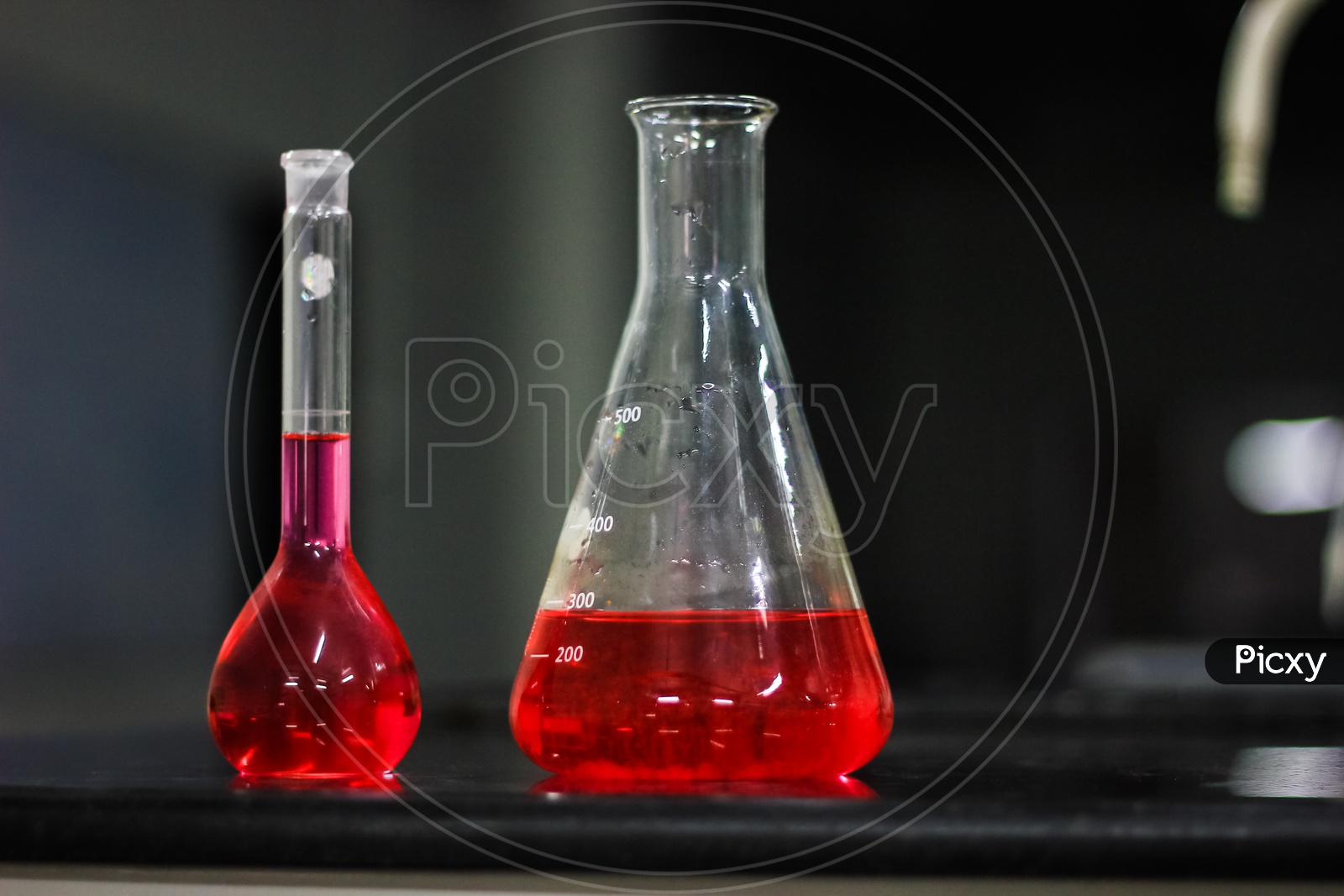 Red Liquid In A Conical Flask And Round Bottomed Flask On A Black Granite Table In Dark Background