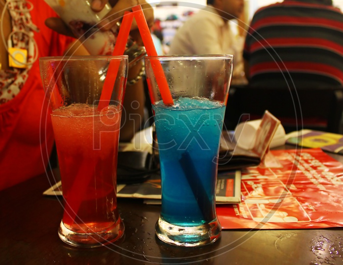 red and blue iced smoothie in long glasses with straws on table