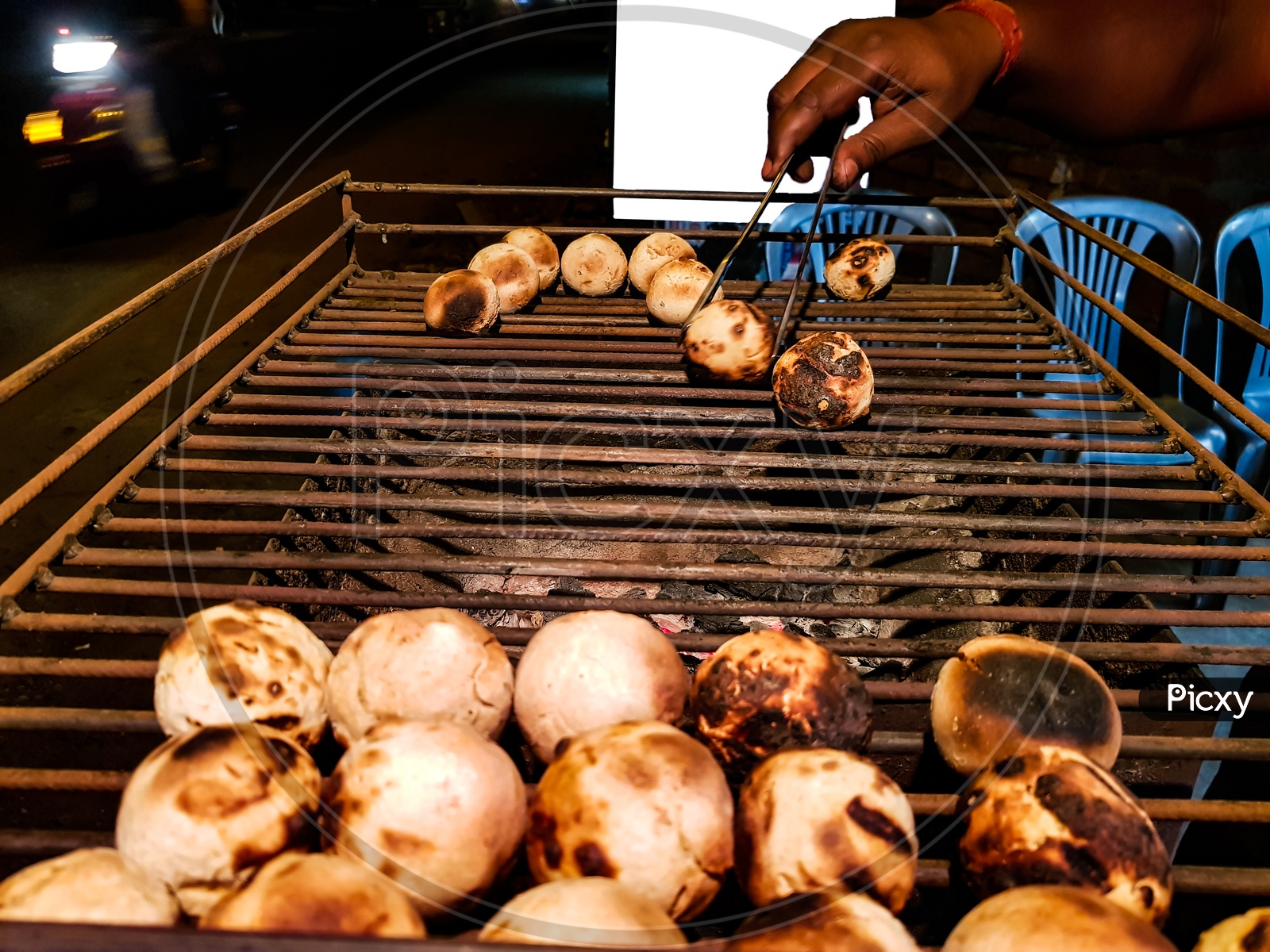 Litti, A Common North Indian Food Is Being Baked Road Side On A Coal Grill By Vendor With White Mock-up Ad Space In The Background