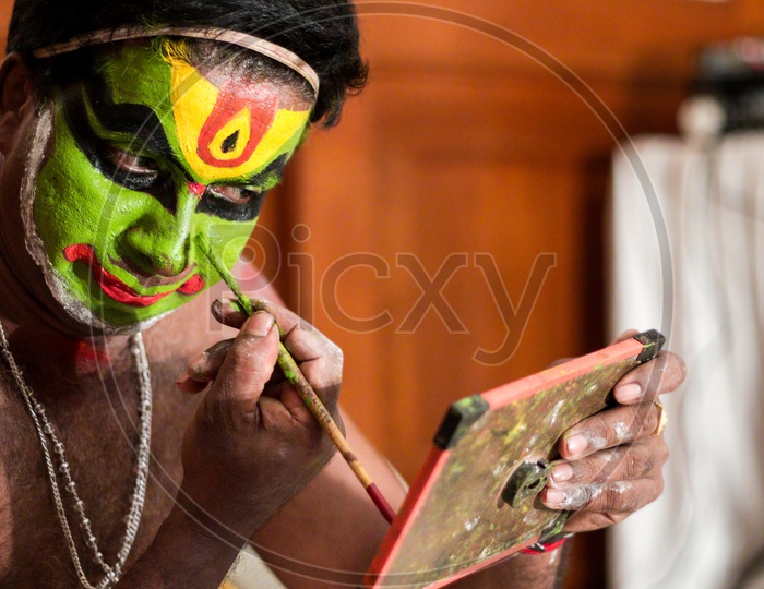 Kathakali dance performer doing face paint and makeup in front of hand held mirror