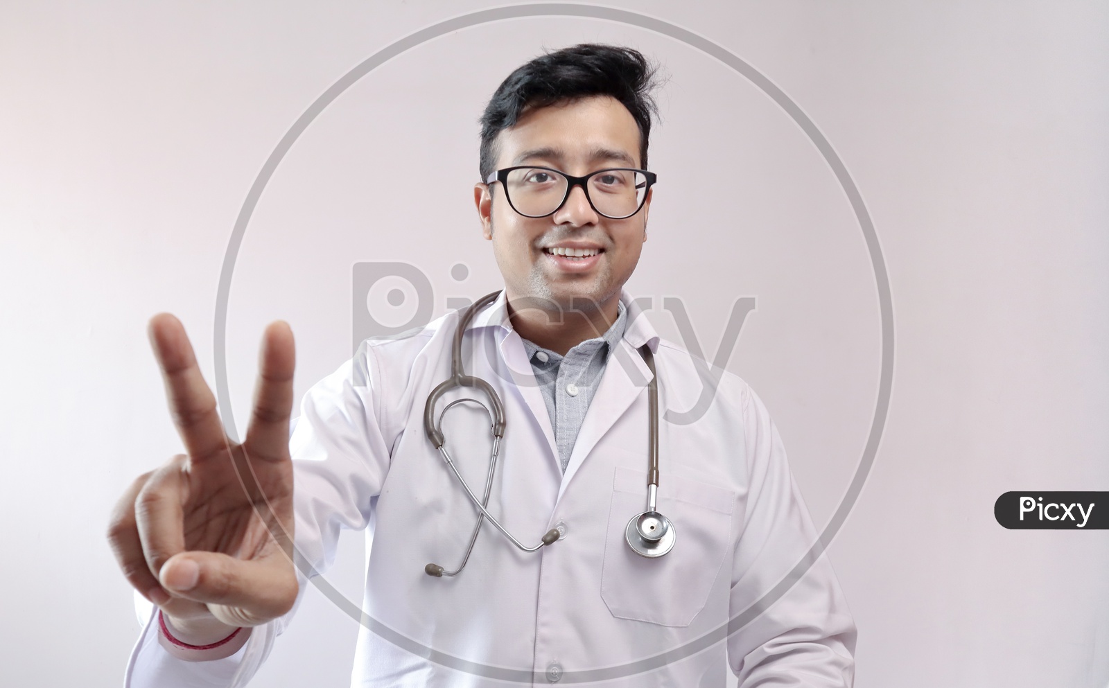 Male Indian Doctor In White Coat And Stethoscope Showing Victory Sign In Confidence