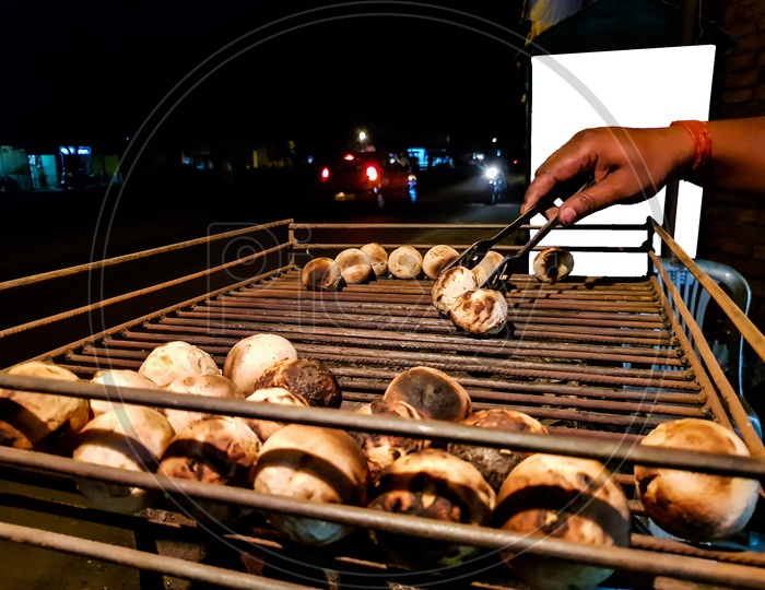 Litti, A Common North Indian Food Is Being Baked Road Side On A Coal Grill By Vendor With White Mock-up Ad Space In The Background