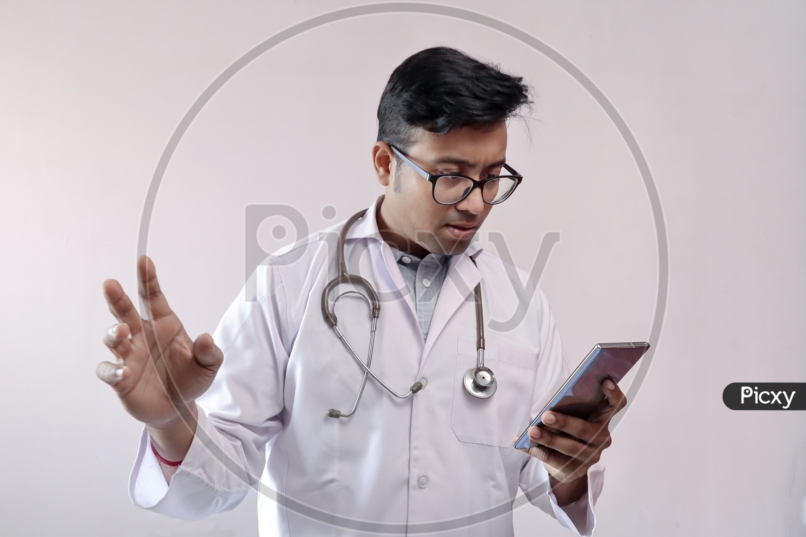 Male Indian Doctor In White Coat And Stethoscope In Worried Expression While Looking At Smart Phone