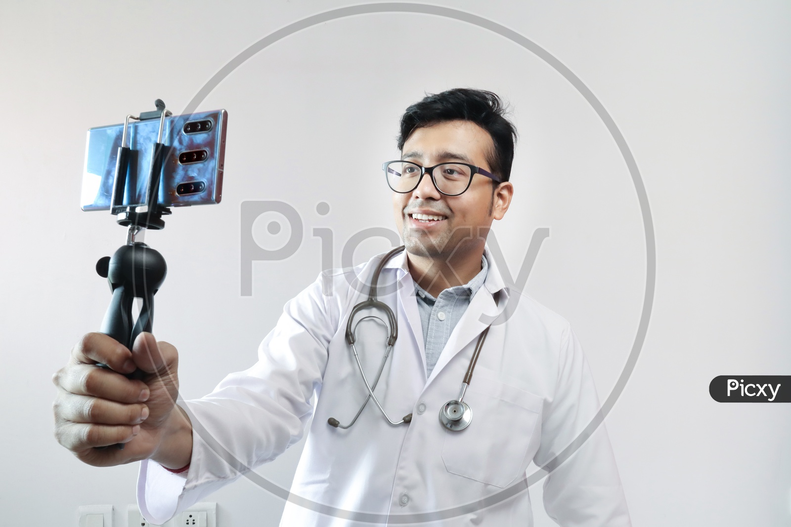 Male Indian Doctor In White Coat And Stethoscope Clicking Selfie With A Mobile Tripod And Futuristic Concept Cameraphone