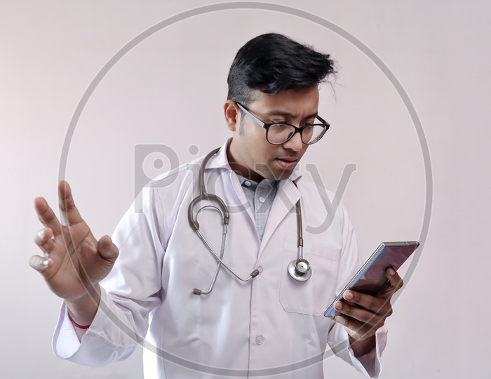 Male Indian Doctor In White Coat And Stethoscope In Worried Expression While Looking At Smart Phone