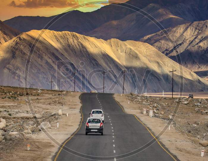 Vehicles on NH1 Road In Leh With Snow Filed Mountains in Background And Sunset Sky