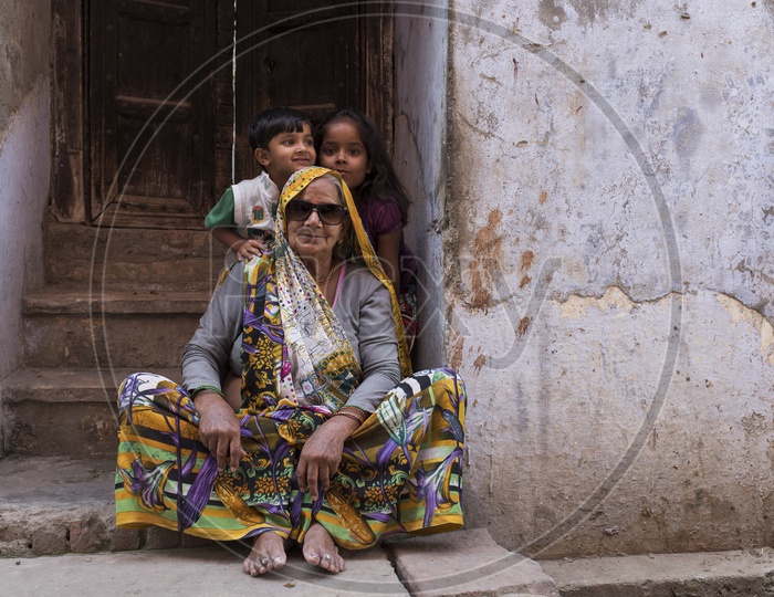 Two children beside an old woman wearing sunglasses sitting by the door