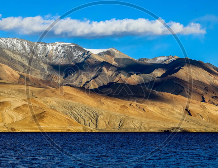 Landscape of snow capped mountains covered with clouds and blue sky alongside the Pangong Lake