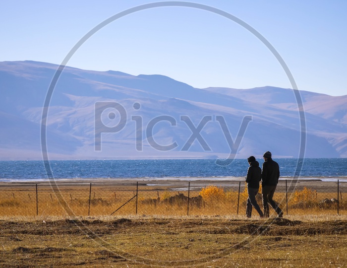 Couple walking alongside the iron fence on the grass lands with mountains and Pangong Lake in the background