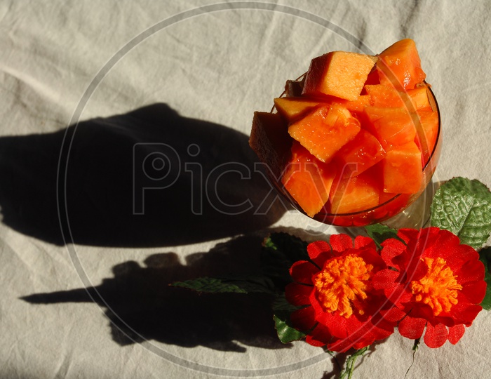 Papaya fruit pieces served in a glass cup with green leaf and red flowers in the background
