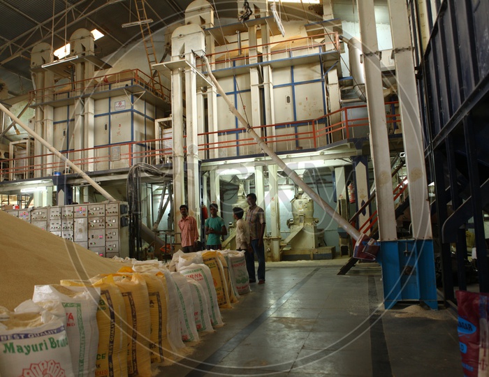 A big rice heap lined with rice bags, machinery and men workers inside a rice mill