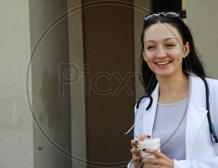 A smiling female doctor with stethoscope with coffee in had wearing a long white coat