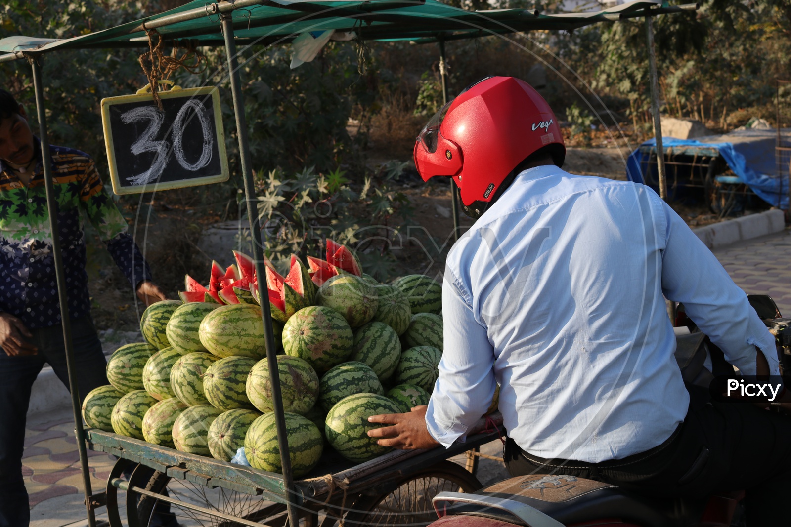 A man wearing helmet sitting on a bike and looking for watermelon to buy