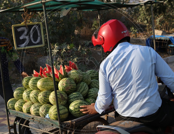 A man wearing helmet sitting on a bike and looking for watermelon to buy