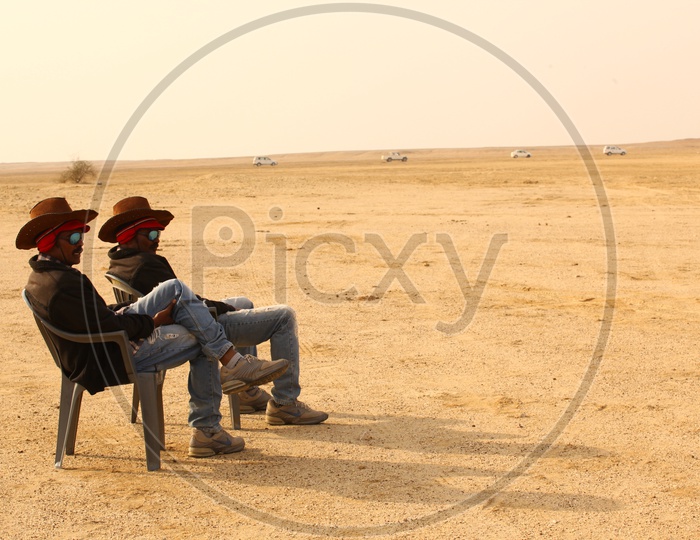 Two men with hats sitting in the chairs at a desert