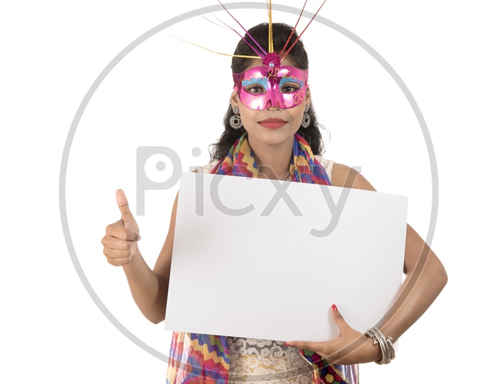 Young Indian Girl Wearing Carnival Mask And Holding White Board With Thump up