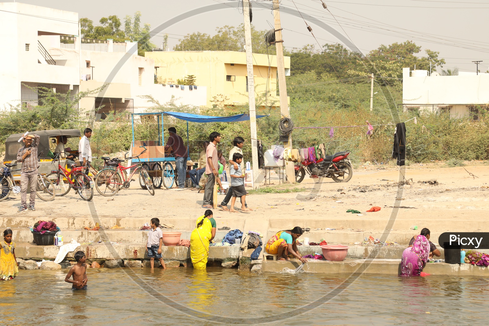 Women washing clothes along the riverside, while kids are playing and men are walking around.