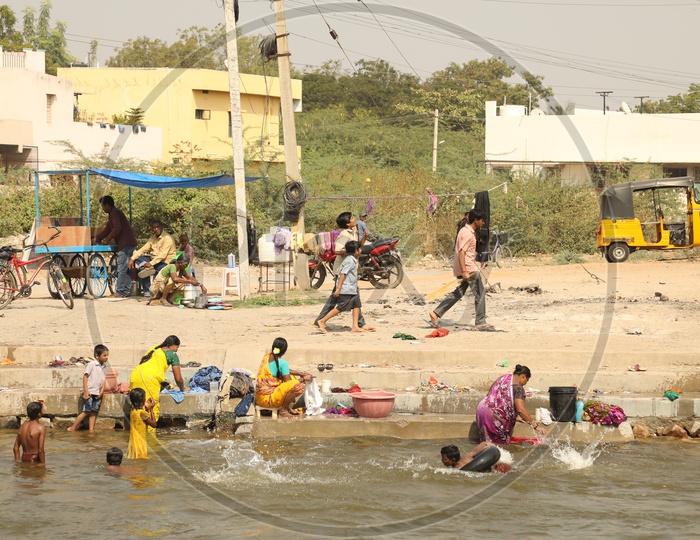Women washing clothes along the riverside, while kids are playing and men are walking around.