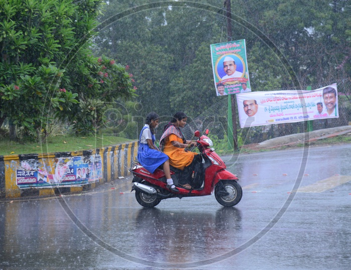 A woman riding a bike with girl behind on the road while its raining