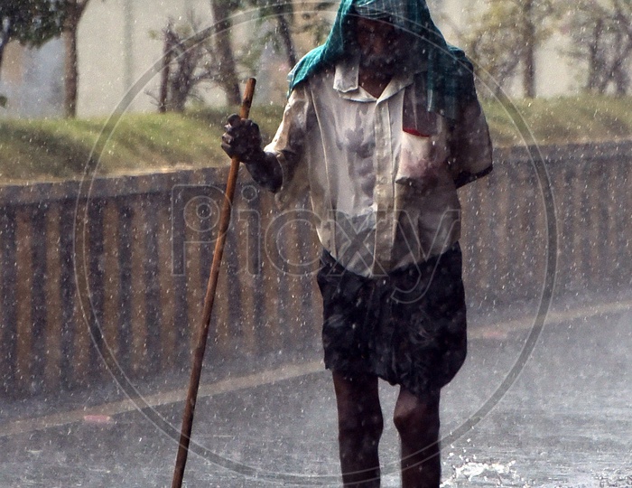 An old man walking on the road with a stick in hand in raini