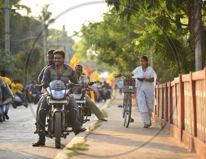 A woman walking on the footpath along with a bicycle as the TDP party supporters were riding bikes on the road during an election campaign rally