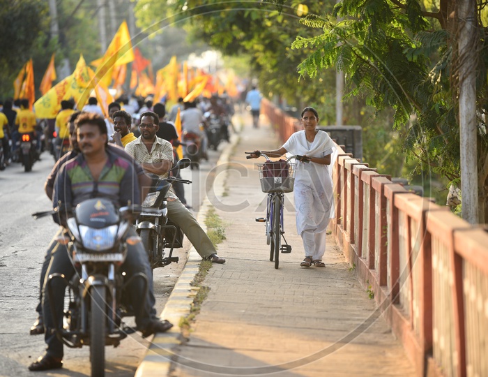 A woman walking on the footpath along with her bicycle as the TDP party supporters were riding bikes on the road during an election campaign rally