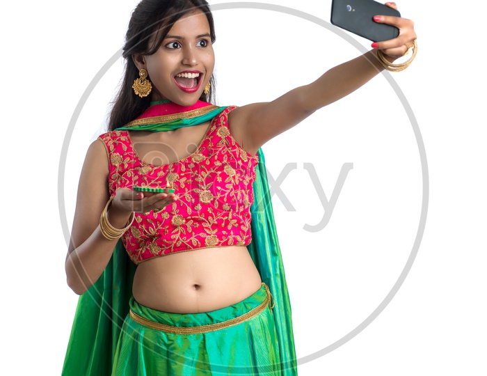 Portrait Of a Young Indian Traditional Girl Holding Festival Dia In Hand And Taking Selfie  in Smart Phone  Over a White Background