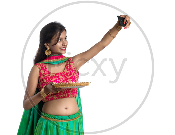 A Young Indian Traditional Girl Taking Selfie In Smart Phone With Dia  or Festival Plate In Hand  Over an Isolated White Background