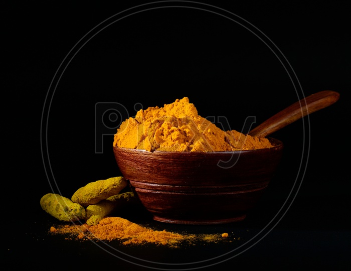 Turmeric powder and roots or sticks in wooden bowl on black background