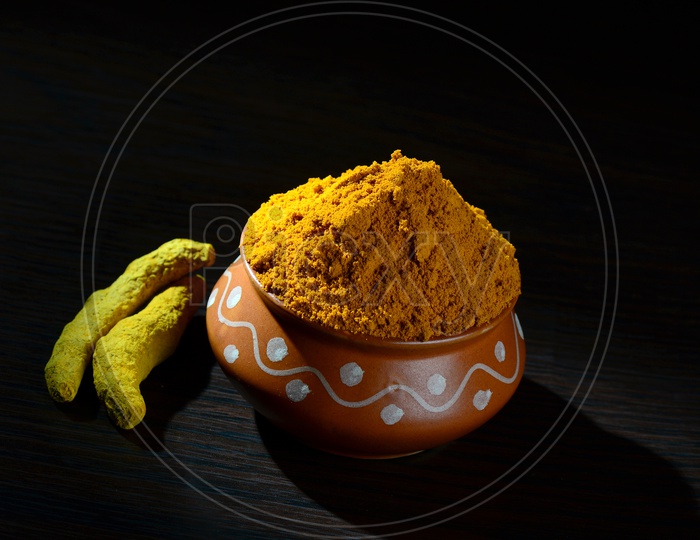 Turmeric powder in clay pot with roots or barks on black background